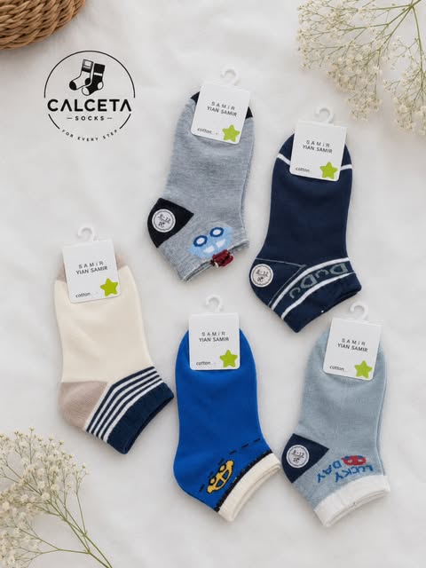 Cinq paires de chaussettes enfant bleu marine, gris, bleu roi et crème, motifs robot, ancre et Lucky Day, disposées à plat sur du lin clair.