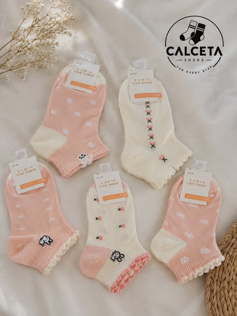 Cinq paires de chaussettes enfant rose poudré et écru, motifs petits moutons, pois et fleurs brodées, volants roses, disposées à plat sur du lin clair.