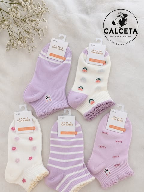 Cinq paires de chaussettes enfant lilas et blanc, motifs cerises, petites fleurs roses, rayures et inscription Luck, disposées à plat sur du lin clair.