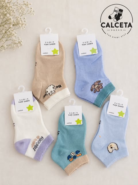 Cinq paires de chaussettes enfant bleu ciel, sable, vert d’eau et écru, motifs chiens et oursons brodés, disposées à plat sur du lin clair.