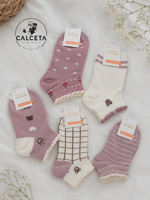 Cinq paires de chaussettes enfant mauve et écru, motifs cœurs, carreaux, rayures et ourson brodé, disposées à plat sur du lin clair.